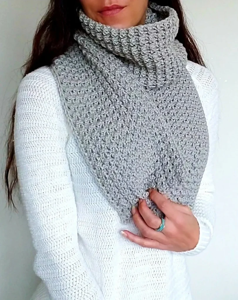 A woman wearing a thick, textured light grey crochet scarf styled as a cozy neck warmer over a white knit sweater. The image showcases the detailed stitch work of the Big Cozy Scarf, a free crochet pattern, highlighting its dense and warm texture.