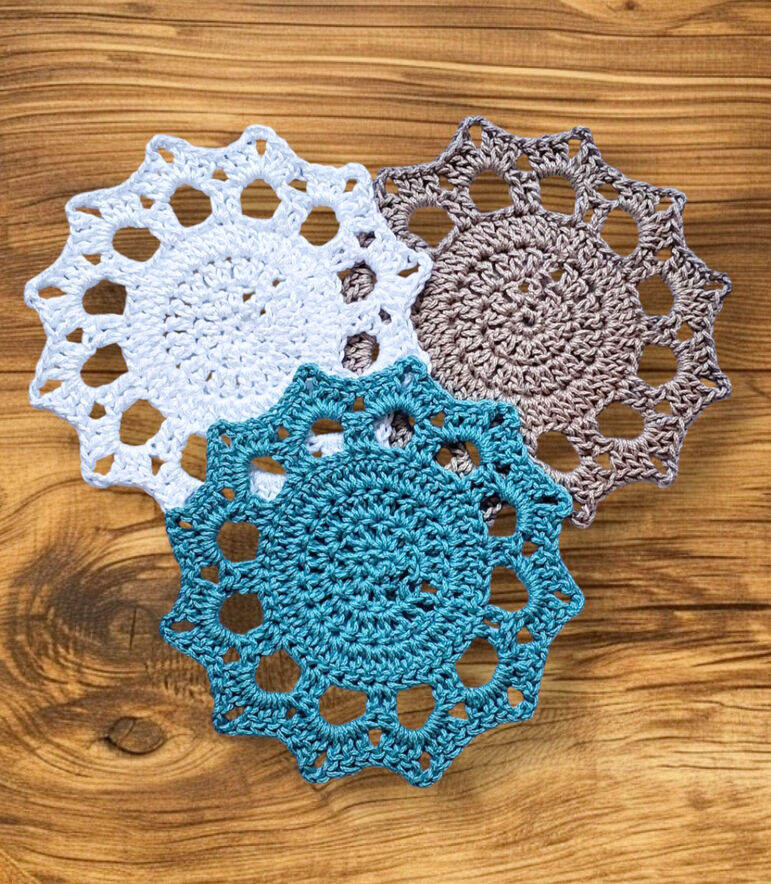 Three Boho Shell Coasters crocheted in white, brown, and teal cotton yarn, arranged on a light wooden background.
