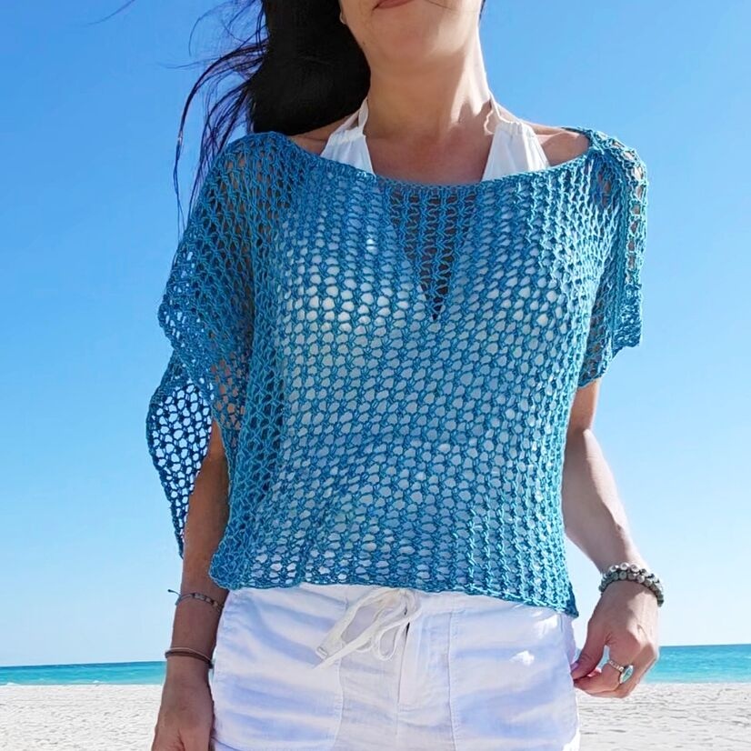 Woman wearing a blue knitted lace poncho beach cover-up on a white sandy beach.