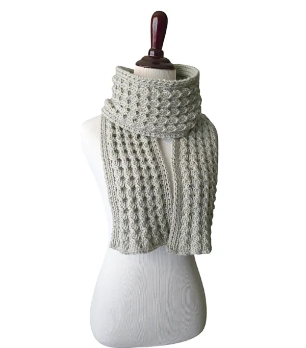 A hand knit scarf modeled on a mannequin against a white background. The stitch design resembles cascading Bells and is made with a light gray yarn.