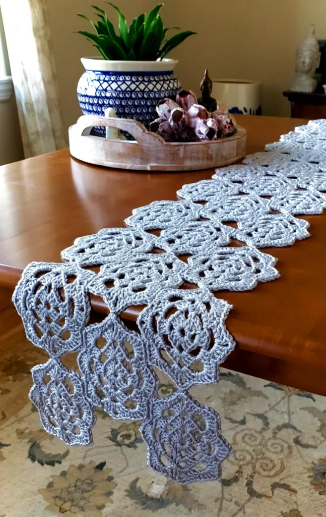 A handmade light gray crochet motif table runner draped over a wooden dining table. The runner features intricate, bohemian lace motifs joined together with a staggered edge. A blue and white ceramic planter sits on a wooden tray in the background, showcasing a coastal boho home decor style.