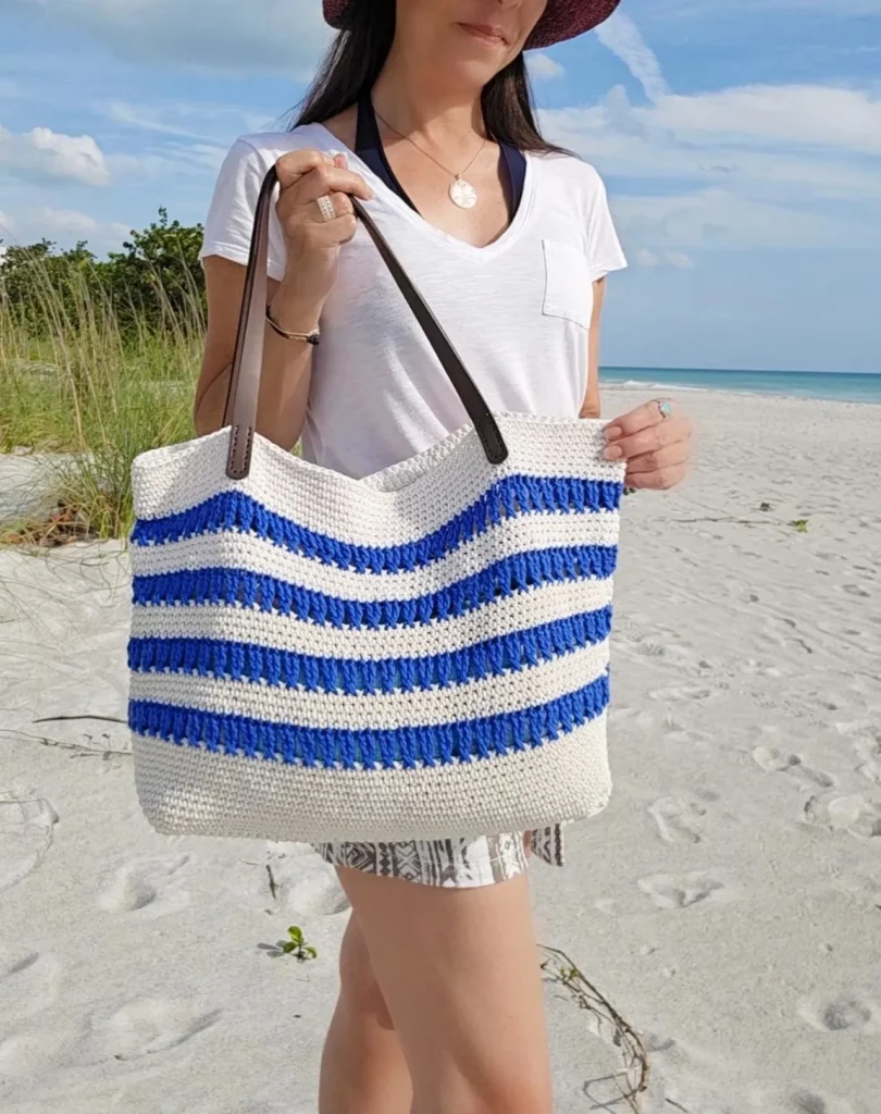 Woman holding a white and blue striped crochet beach tote with leather handles on a sunny beach.