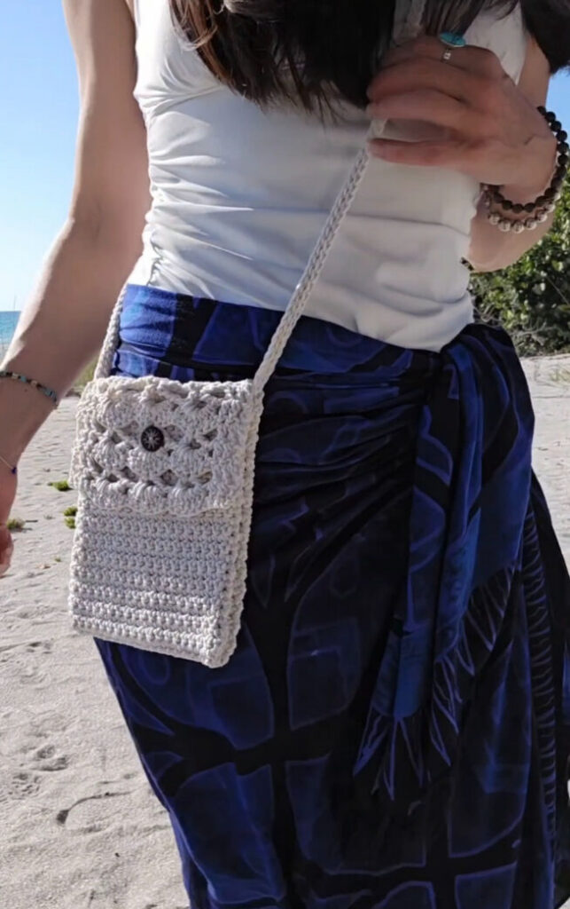 A woman wearing a handmade crochet crossbody bag on a sunny beach.