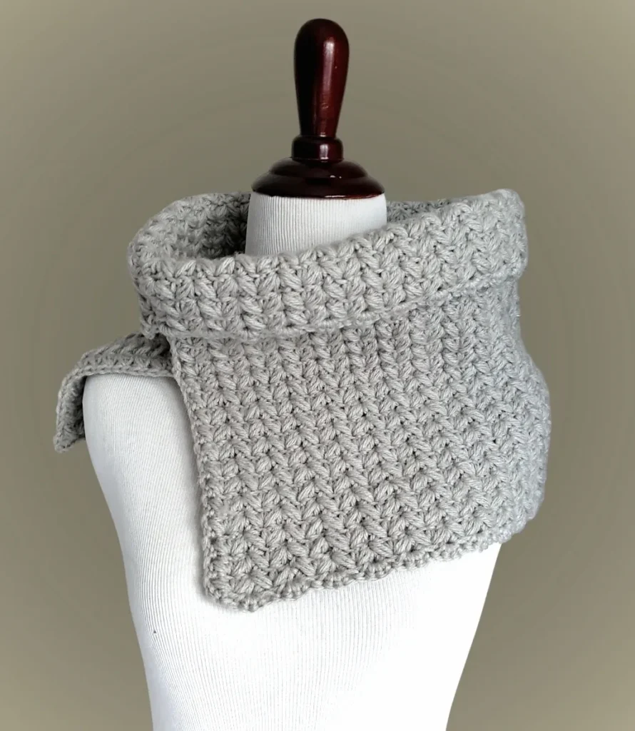 A cozy, handmade cowl in a cream colored yarn, featuring the textured Dapper Puff Stitch. It is displayed on a mannequin highlighting the intricate details.