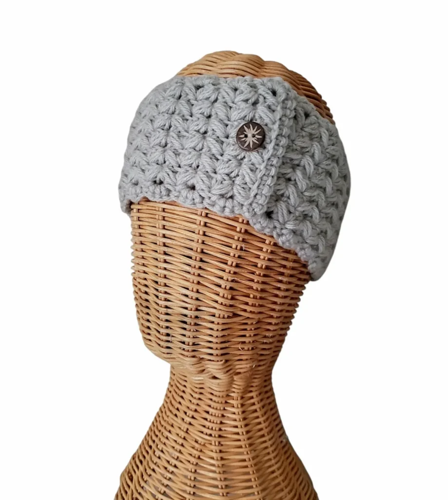 A free crochet pattern for the Dapper Puff Stitch shown as a headband. It is modeled on a wooden mannequin and is made with a cream colored yarn.
