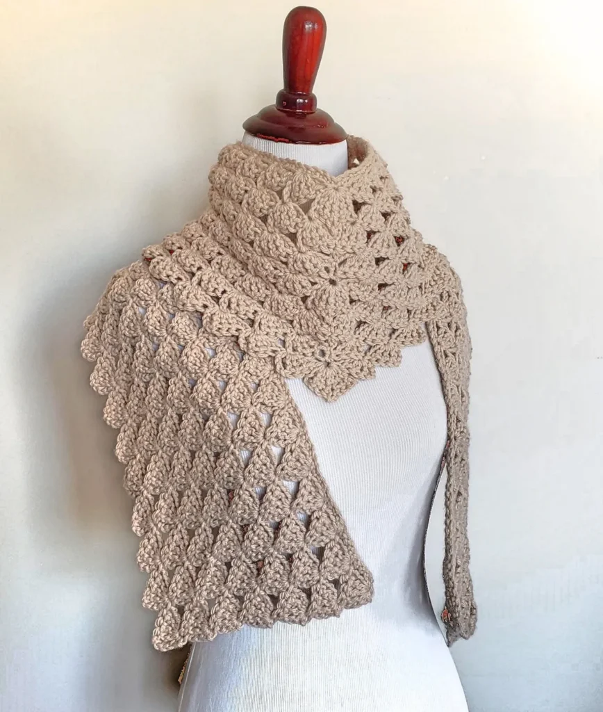 Cream-colored crocheted scarf with a shell stitch pattern displayed on a white mannequin torso, showcasing the elegant drape and texture of the Drifting Shells Scarf.