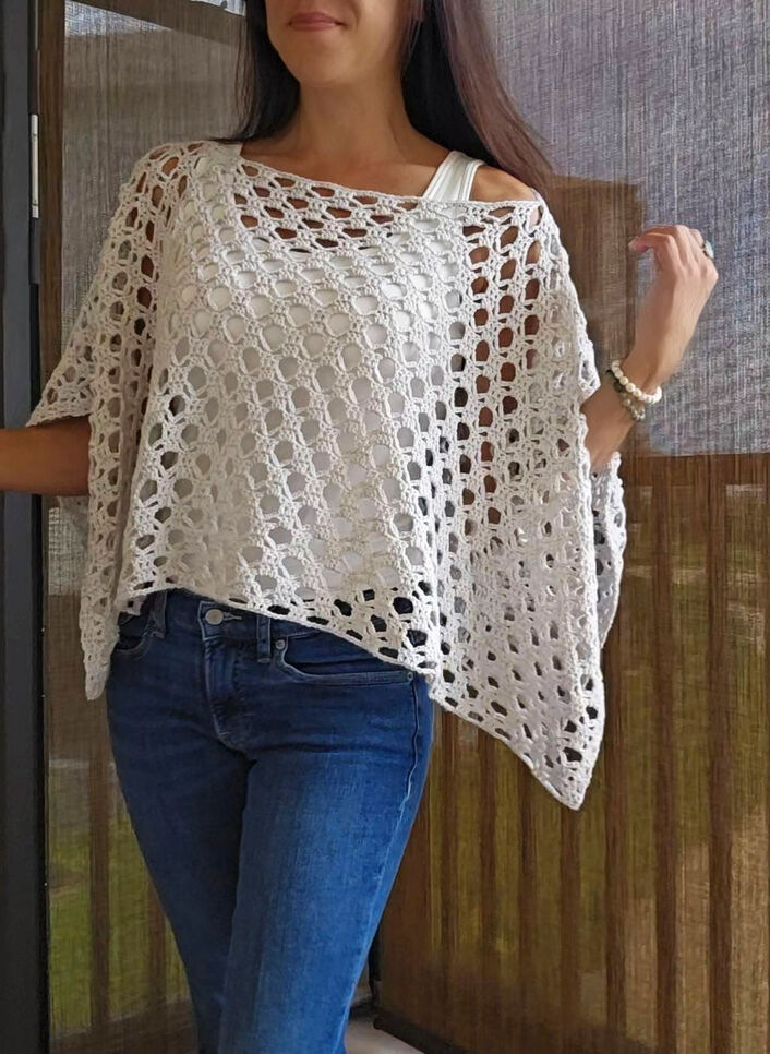 A woman wearing a lightweight, cream-colored openwork crochet poncho over a white tank top and blue jeans.
