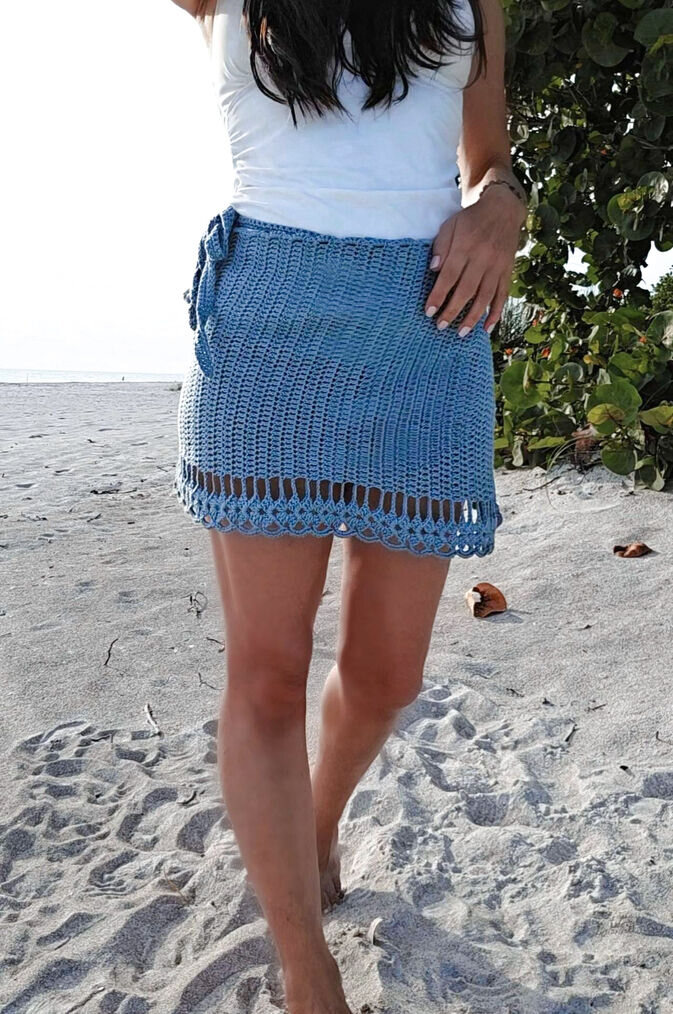 A woman on a sandy beach wearing a light blue crochet wrap skirt.The sarong features a solid stitch body and an open work hanging shell for the bottom hem.