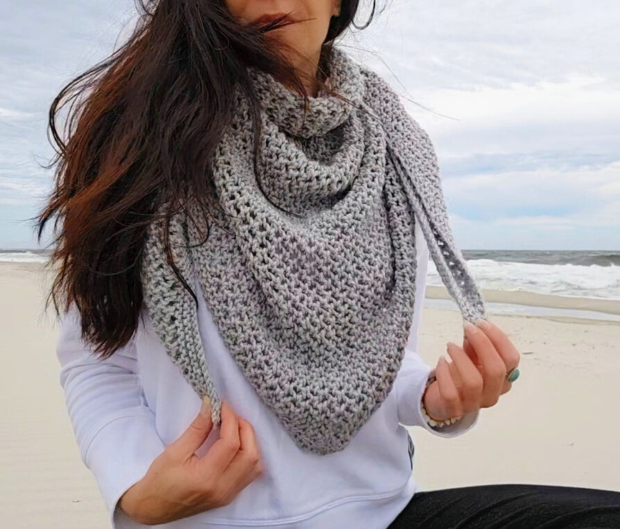 Close-up of a textured grey knit triangular shawl with eyelet details, worn by a woman on a sandy beach.