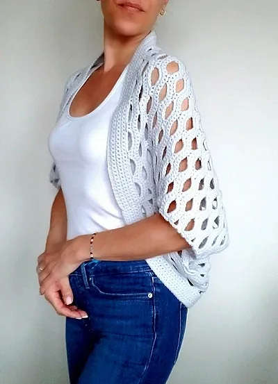 A woman from the side profile wearing a light grey crocheted cocoon cardigan featuring a prominent openwork shell stitch pattern with large teardrop-shaped gaps. She is pairing it with a white tank top and blue jeans against a plain white background.