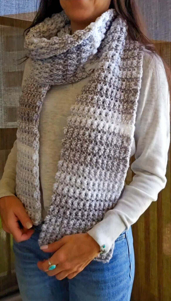 A woman models a long, textured crochet scarf in gradient shades of white, grey, and taupe.