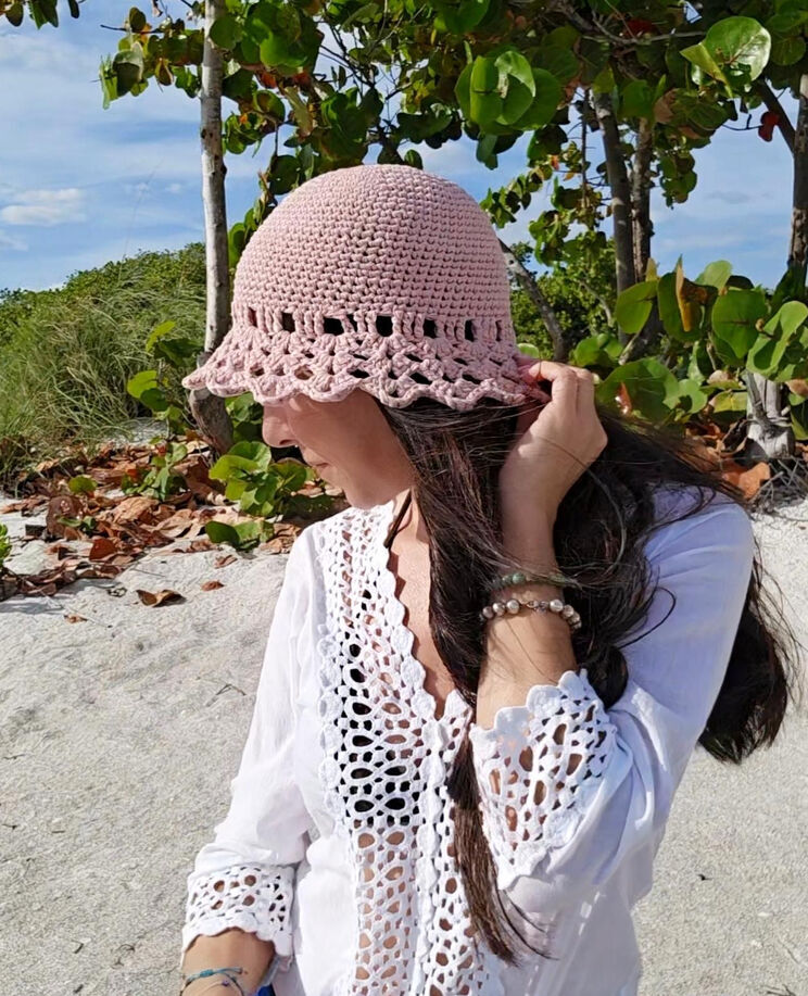 A free crochet pattern for a summer sun hat in a light pink color styled on a model sitting on the beach.